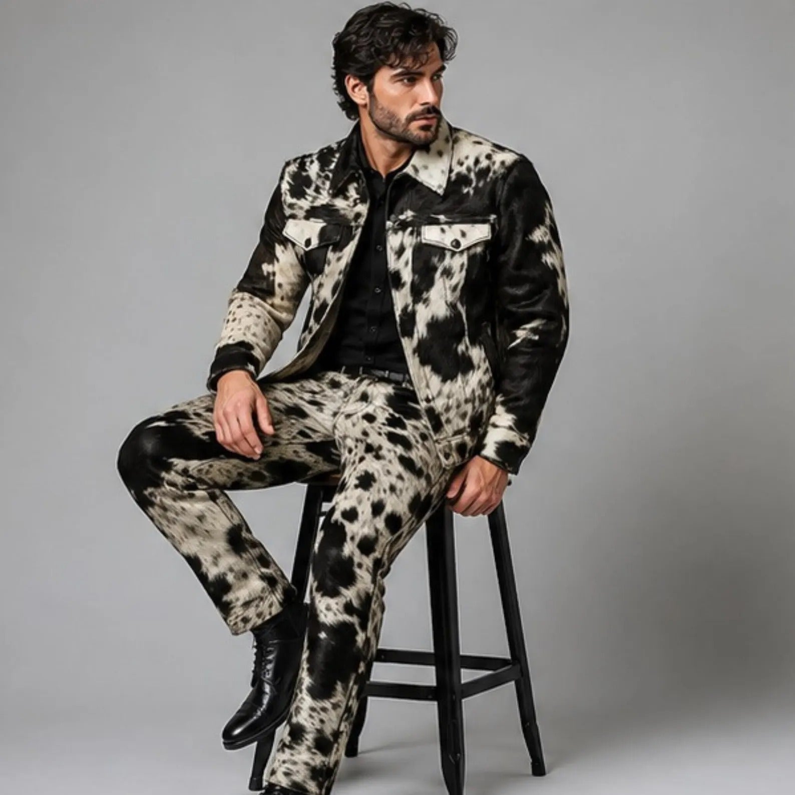 A man wearing a black and beige cow-print leather jacket and matching pants, sitting on a black stool against a gray studio background.