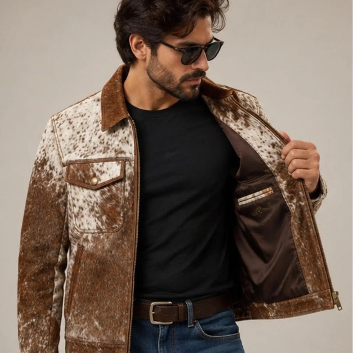 A man wearing a brown and white cow-print leather jacket with a brown suede collar, shown wearing a black t-shirt and blue jeans against a neutral studio background.